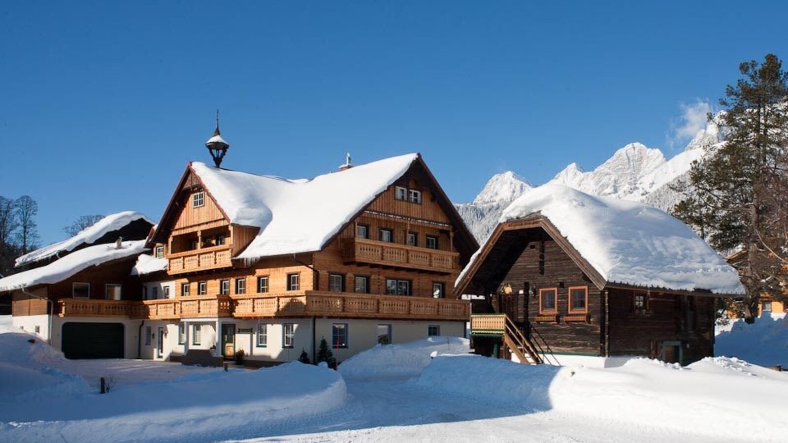 The Farm House and a smaller wooden outbuilding, both covered in snow, with snow-capped mountains visible in the distance.