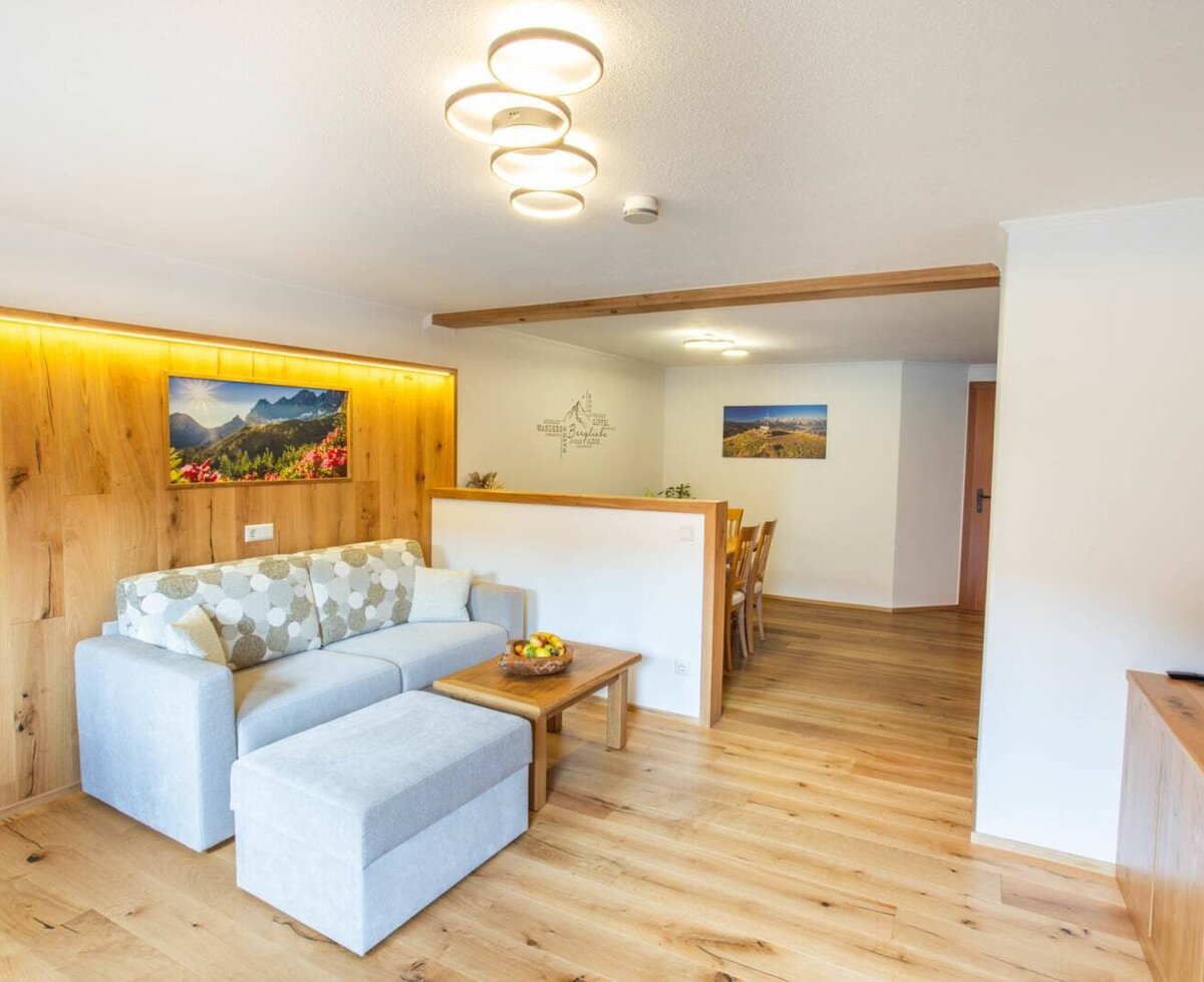 The living area of the farm house features a sofa, ottoman, coffee table, and a wooden accent wall, connected to a dining area with a wooden table and chairs.