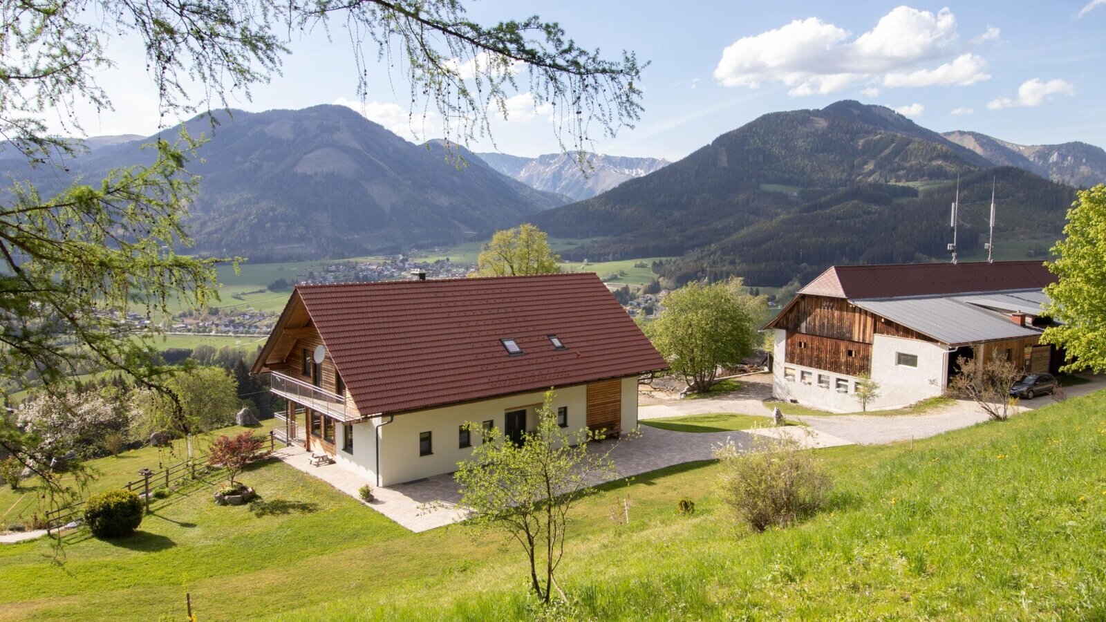 The farmhouse with a balcony and an adjacent farm building, situated on a green hillside with views of the surrounding mountains and valley.