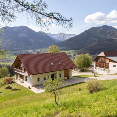 The farmhouse with a balcony and an adjacent farm building, situated on a green hillside with views of the surrounding mountains and valley.