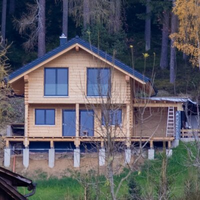 The exterior of the wooden farm house, featuring a dark roof, multiple windows, and an elevated foundation, is set against a dense forest.