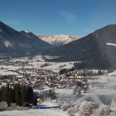 A snow-covered village nestled in the valley surrounded by mountains, featuring ski slopes with active snow cannons for winter activities near the farmhouse.