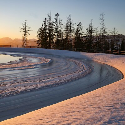 View from the Farm House over a snowy landscape with a partially frozen pond, distant mountains, and trees illuminated by the dawn or dusk light.