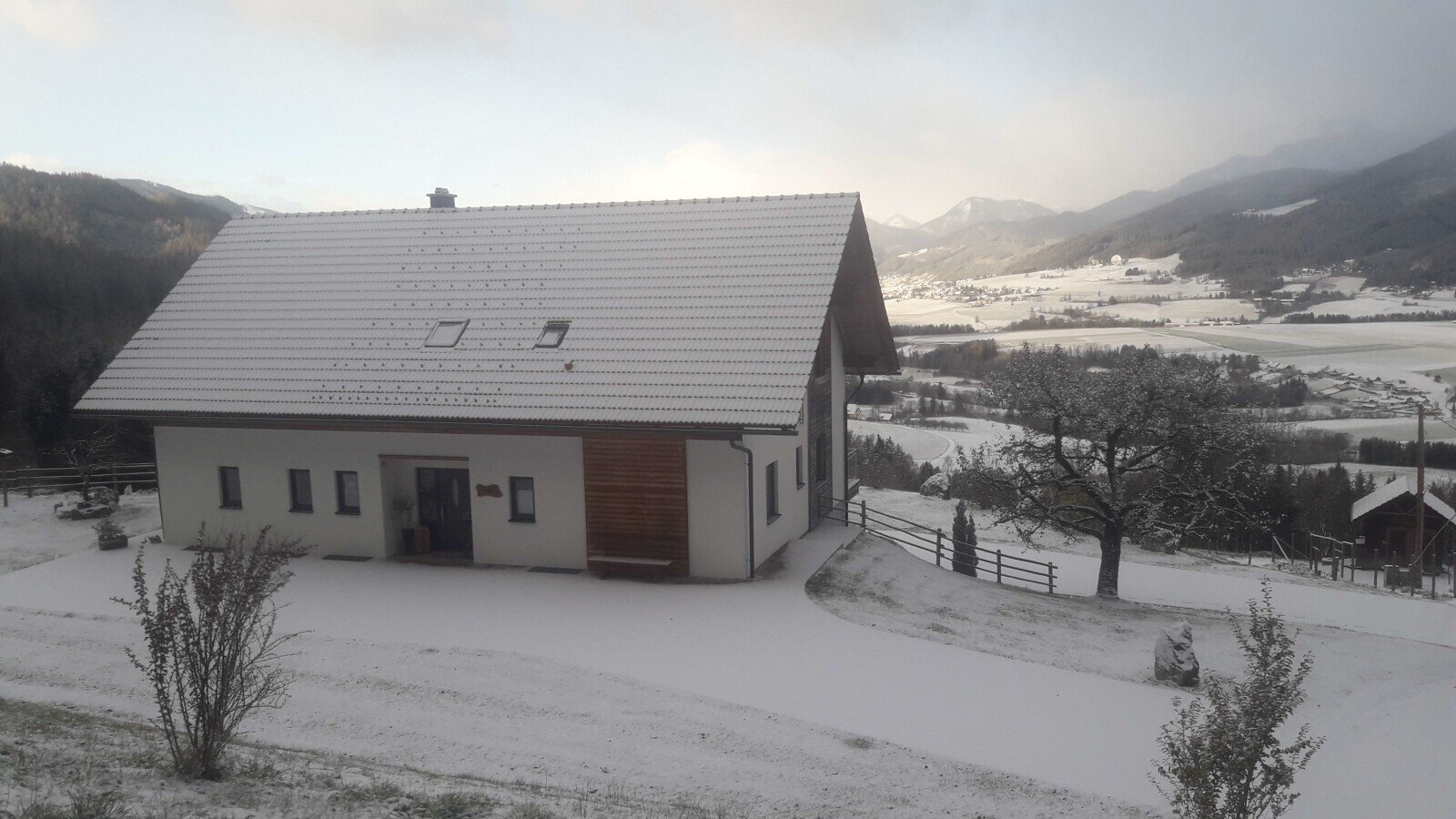 The snow-covered farmhouse, with its white exterior and gabled roof, overlooks a winter valley landscape with distant mountains.