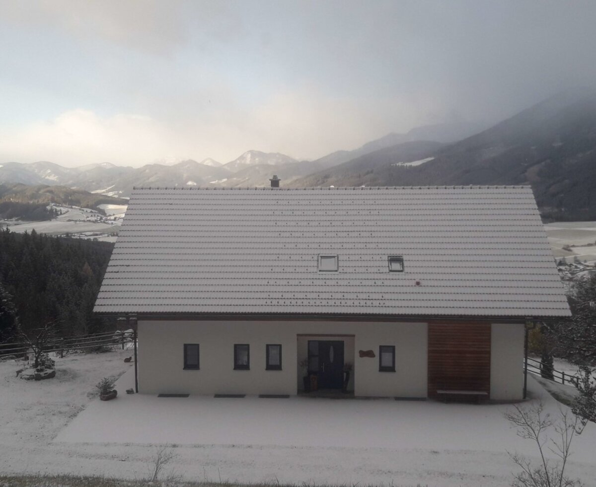 The snow-covered farmhouse exterior features a white facade and a gray tiled roof, set against a mountain and valley landscape.