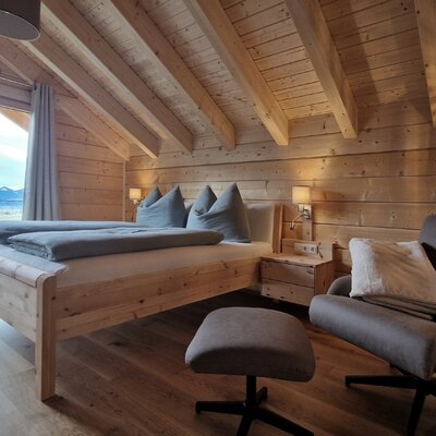 The Farm House bedroom features a wooden bed, an armchair with an ottoman, and a floor-to-ceiling glass door providing mountain views.