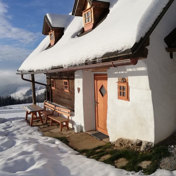 The farmhouse exterior, featuring a snow-covered roof, an entrance door, and outdoor wooden benches with a table, set against a snowy mountain backdrop.