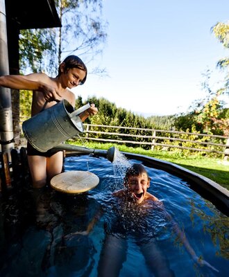 An outdoor wooden bathing tub at the farmhouse, with guests using a watering can for showering.