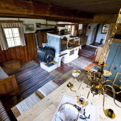 The farmhouse's kitchen/living room, featuring a wood-burning stove, a tiled stove, and a dining area with wooden benches.