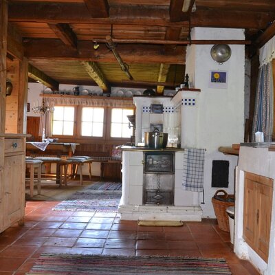 The kitchen-living room of the mountain cabin, featuring a wood stove with a tiled oven and traditional wooden furnishings.