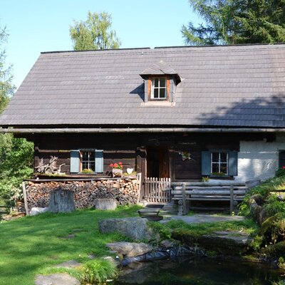 The mountain cabin with a wooden facade, shingle roof, a stack of firewood, and a small pond in the foreground.