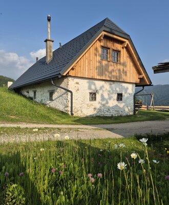 The farmhouse exterior, featuring a stone base, wooden upper facade, dark roof, and a visible chimney.