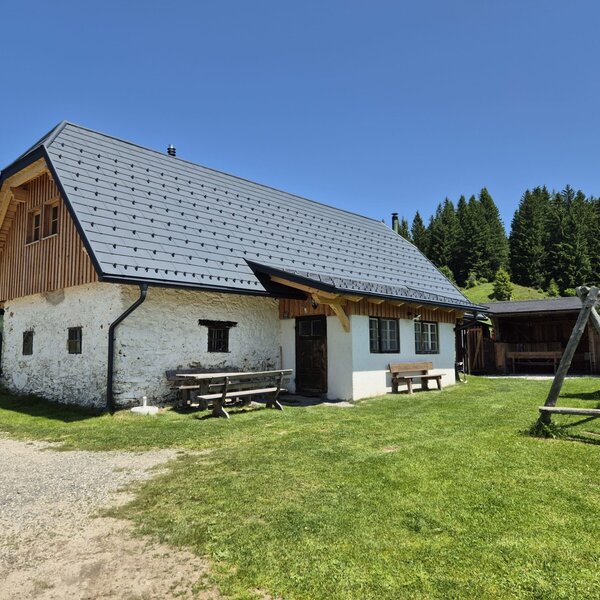 The farmhouse exterior, featuring white stone walls, a grey roof, outdoor benches, a covered seating area, and a wooden swing set.