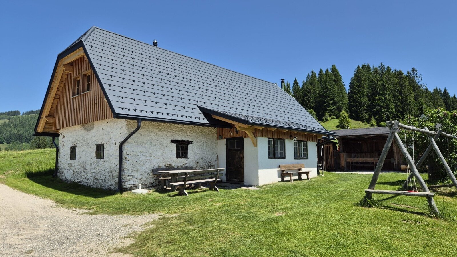 The farmhouse exterior, featuring white stone walls, a grey roof, outdoor benches, a covered seating area, and a wooden swing set.