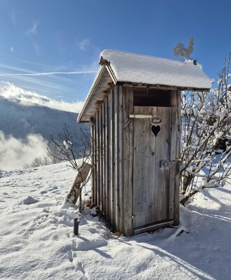 A snow-covered wooden outhouse at the farmhouse, featuring a heart-shaped cutout on the door and a rooster weathervane.