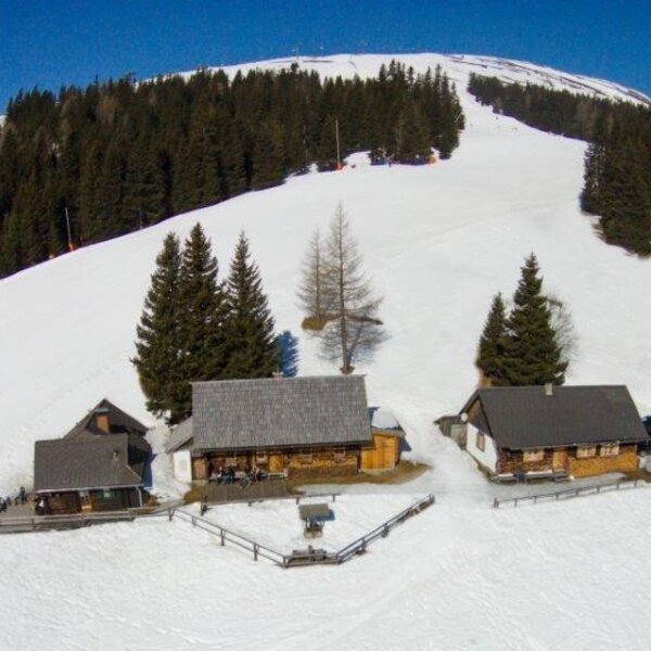 The farmhouse is located directly on a snow-covered mountain ski slope, surrounded by pine trees and adjacent buildings.
