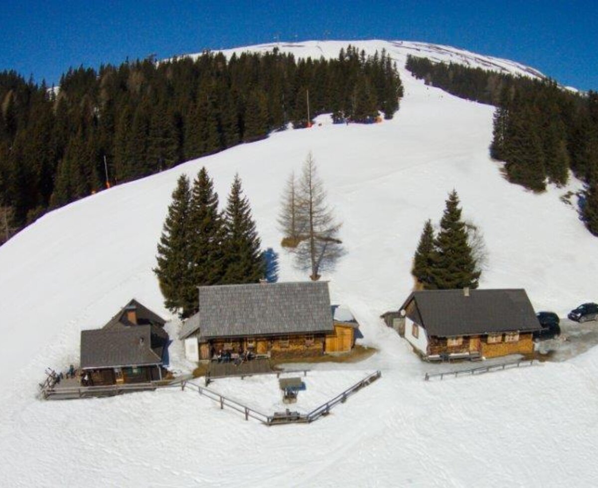 The farmhouse is located directly on a snow-covered mountain ski slope, surrounded by pine trees and adjacent buildings.