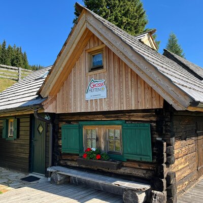 The exterior of the farmhouse, featuring rustic wooden construction, green window shutters, and flower boxes, surrounded by a grassy landscape.