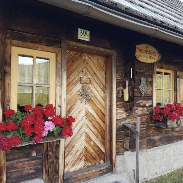 The entrance of the farmhouse features a wooden door with house number 394, windows with yellow frames and red geranium flower boxes, and a traditional wooden exterior.