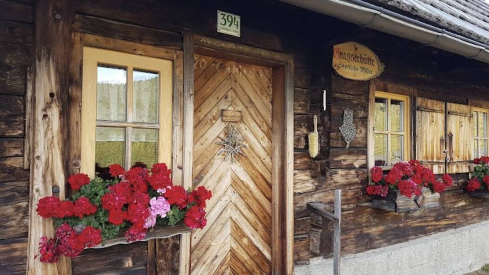 The entrance of the farmhouse features a wooden door with house number 394, windows with yellow frames and red geranium flower boxes, and a traditional wooden exterior.