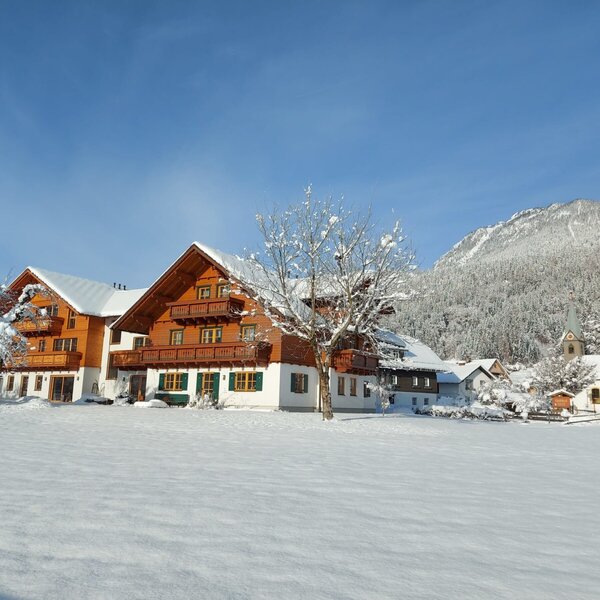 The Farm House exterior in winter, featuring traditional wooden architecture, snow-covered grounds, and distant mountains with a small chapel.