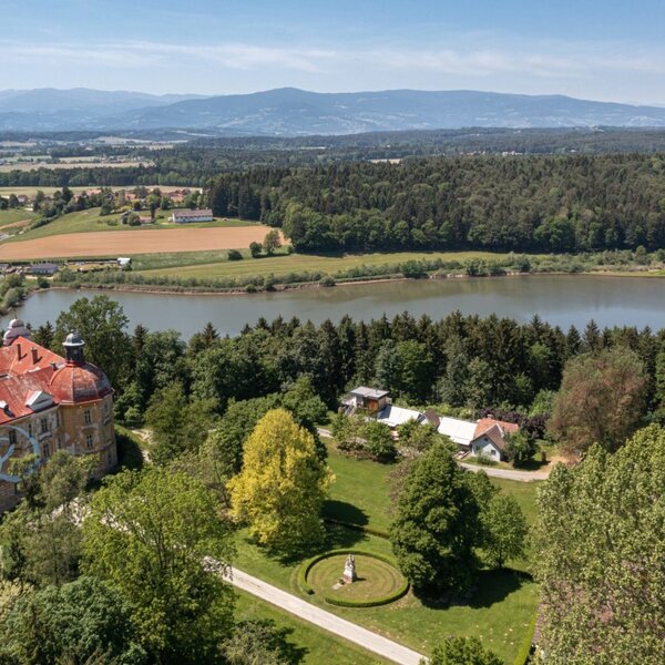 Scenic aerial view Gut Hornegg surrounded by expansive green fields and forests, overlooking an idyllic lake.