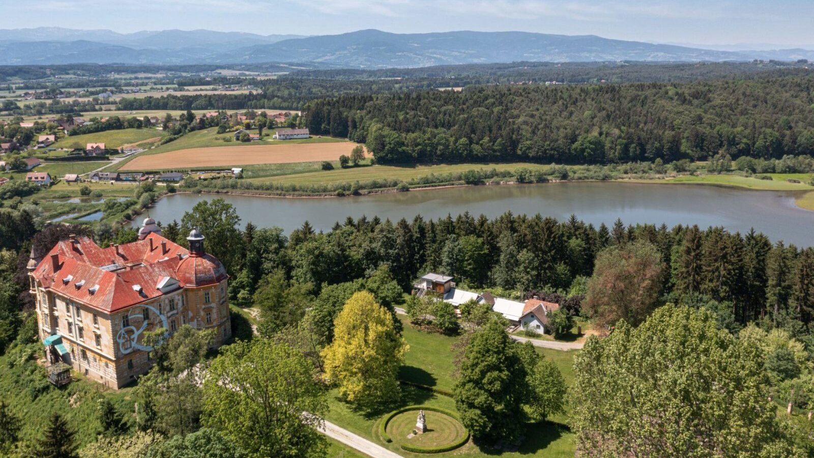 Scenic aerial view Gut Hornegg surrounded by expansive green fields and forests, overlooking an idyllic lake.