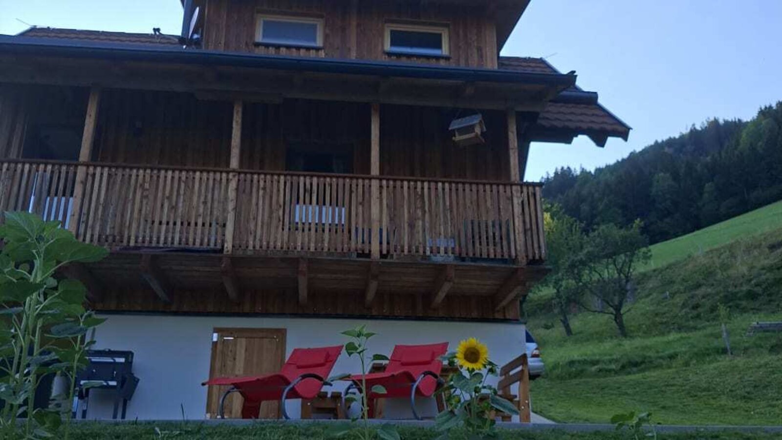 The inviting wooden bed and breakfast featuring comfortable red lounge chairs on the lawn against a scenic green hillside.