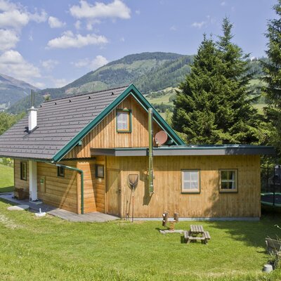 The wooden farmhouse, surrounded by a lawn, includes a small picnic area and a trampoline, with mountains visible in the background.