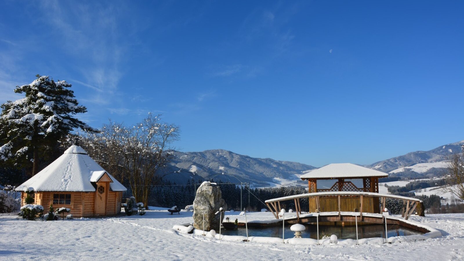 The Farm House grounds feature a snow-covered wooden hut and a gazebo with a bridge over a pond, set against a backdrop of snowy mountains.