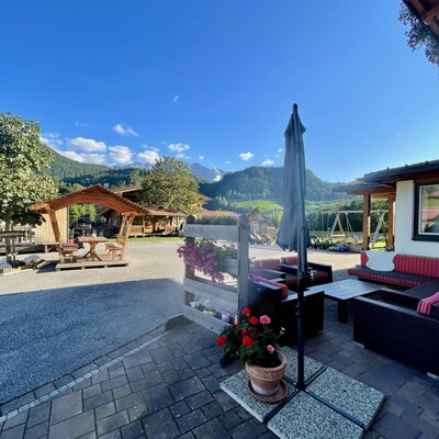 Paved terrace of the farmhouse with outdoor seating, an umbrella, mountain views, and a playground in the background.