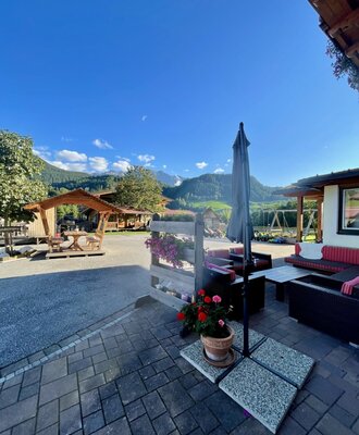 Paved terrace of the farmhouse with outdoor seating, an umbrella, mountain views, and a playground in the background.