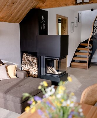 The living area of the farm house features a modern corner fireplace with firewood, integrated storage, a wooden staircase, and exposed ceiling beams.