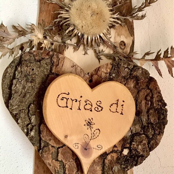 A wooden welcome decoration in the Farm House, featuring a heart with bark, a dried thistle, and the greeting "Grias di" carved into it.