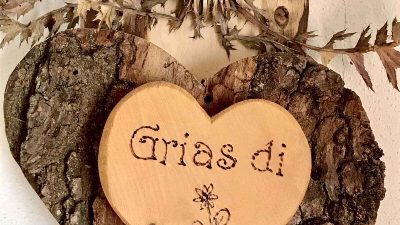A wooden welcome decoration in the Farm House, featuring a heart with bark, a dried thistle, and the greeting "Grias di" carved into it.