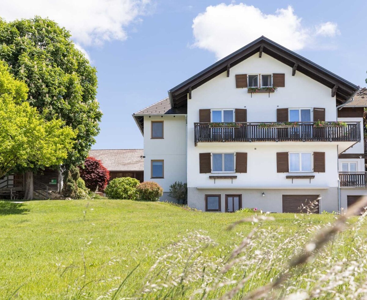 The farmhouse's inviting exterior featuring a lush green lawn and a large shade-providing tree under a clear blue sky.
