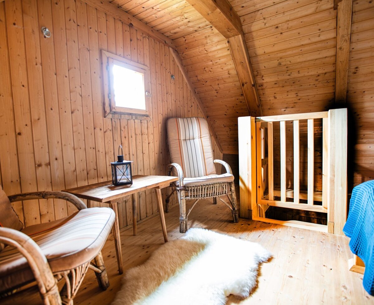 A wooden-paneled attic room in the farmhouse features two armchairs, a small table, and a wooden safety gate.