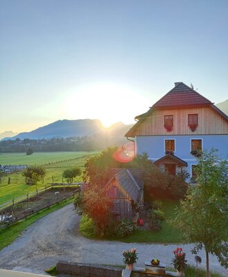 The Farm House exterior, set against mountains and green fields, features a wooden upper story, a blue facade, a gravel path, and a foreground water feature at sunrise.