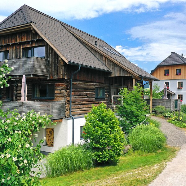 The farm house exterior, featuring traditional wooden log construction, multiple balconies, and surrounding greenery with a gravel path.