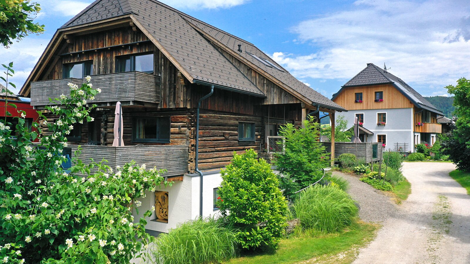 The farm house exterior, featuring traditional wooden log construction, multiple balconies, and surrounding greenery with a gravel path.