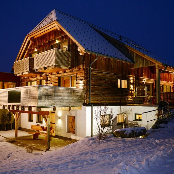The illuminated wooden Farm House exterior at night, featuring multiple balconies and a snow-covered environment.