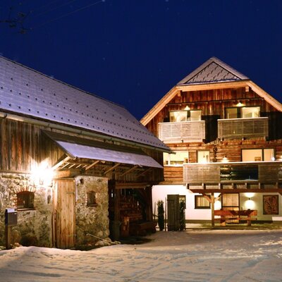The snow-covered exterior of the Farm House at night, featuring the main building with illuminated balconies and an adjacent traditional barn.