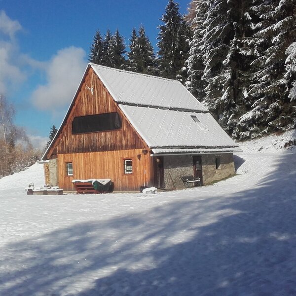 The farmhouse exterior in a snowy winter setting, featuring wooden and stone construction and surrounded by snow-dusted pine trees. The farmhouse exterior in a snowy winter setting, featuring wooden and stone construction and surrounded by snow-dusted pine trees.