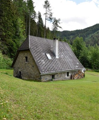 Exterior view of the cabin with stone walls, a tiled roof, chimney, and a stack of firewood, surrounded by grass and forest.