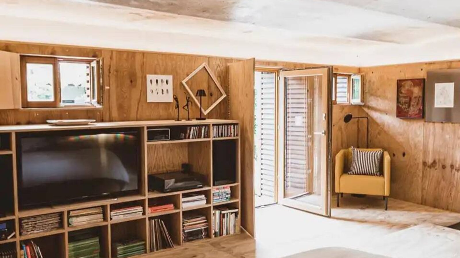 Bedroom in the farmhouse with a large bed, wooden walls, a shelving unit with a TV and books, a yellow armchair, and outdoor access.