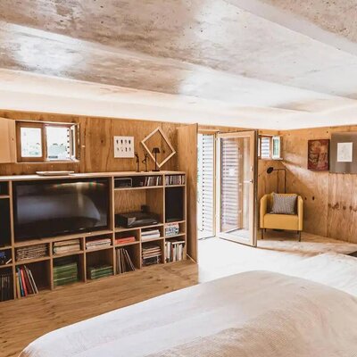 Bedroom in the farmhouse with a large bed, wooden walls, a shelving unit with a TV and books, a yellow armchair, and outdoor access.