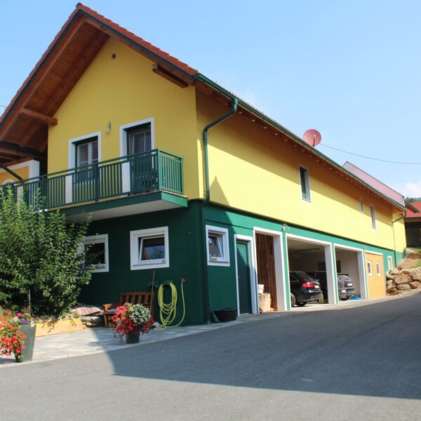 The yellow and green exterior of the farm house, featuring a balcony, ground-floor garages, and a small garden area.