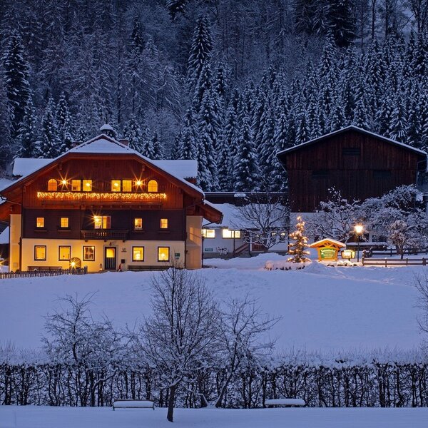 The farm house exterior, illuminated with warm lights, set against a snow-covered landscape and a dense forest backdrop. The farm house exterior, illuminated with warm lights, set against a snow-covered landscape and a dense forest backdrop.