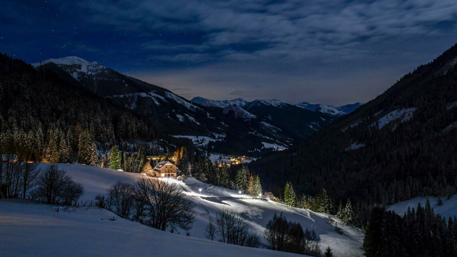 The brightly lit farmhouse nestled in an idyllic, snow-covered mountain landscape under a starry night sky.