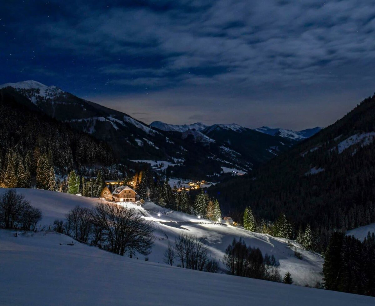 The brightly lit farmhouse nestled in an idyllic, snow-covered mountain landscape under a starry night sky.